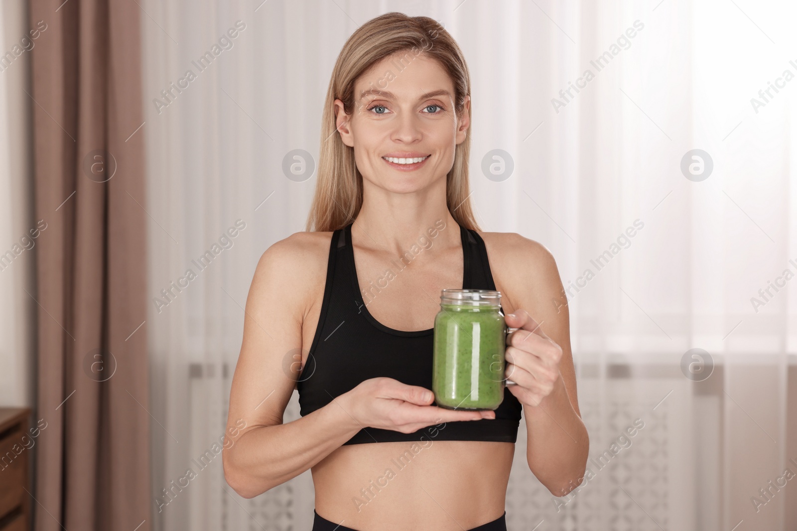 Photo of Young woman in sportswear with mason jar of fresh smoothie at home