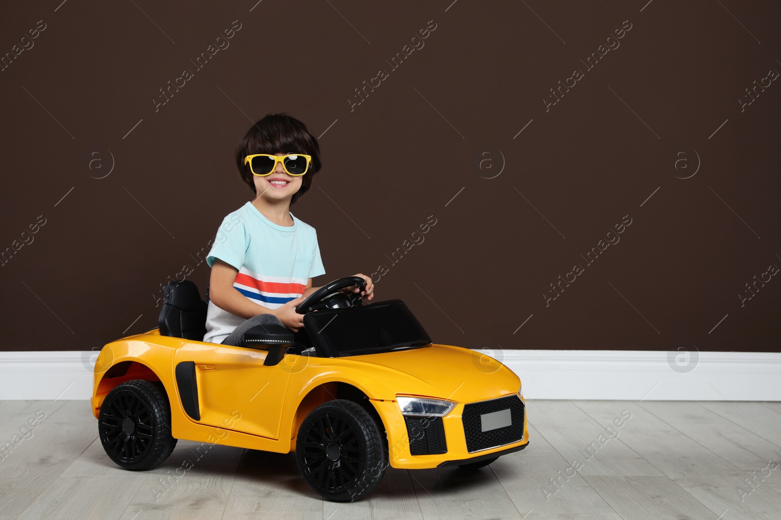 Photo of Cute little boy driving children's electric toy car near brown wall indoors. Space for text