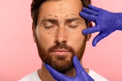 Doctor checking patient's face before cosmetic surgery operation on pink background, closeup