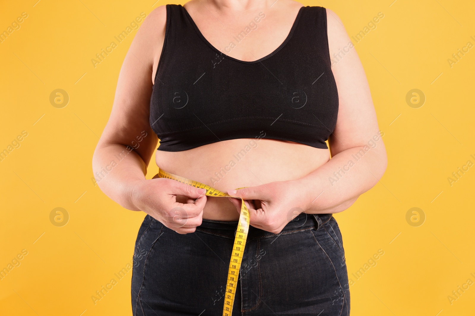 Photo of Overweight woman measuring waist with tape on yellow background, closeup