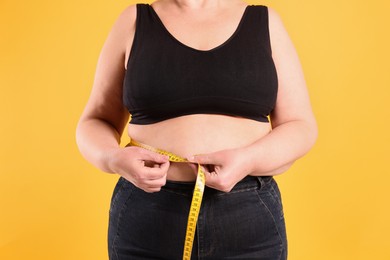 Overweight woman measuring waist with tape on yellow background, closeup