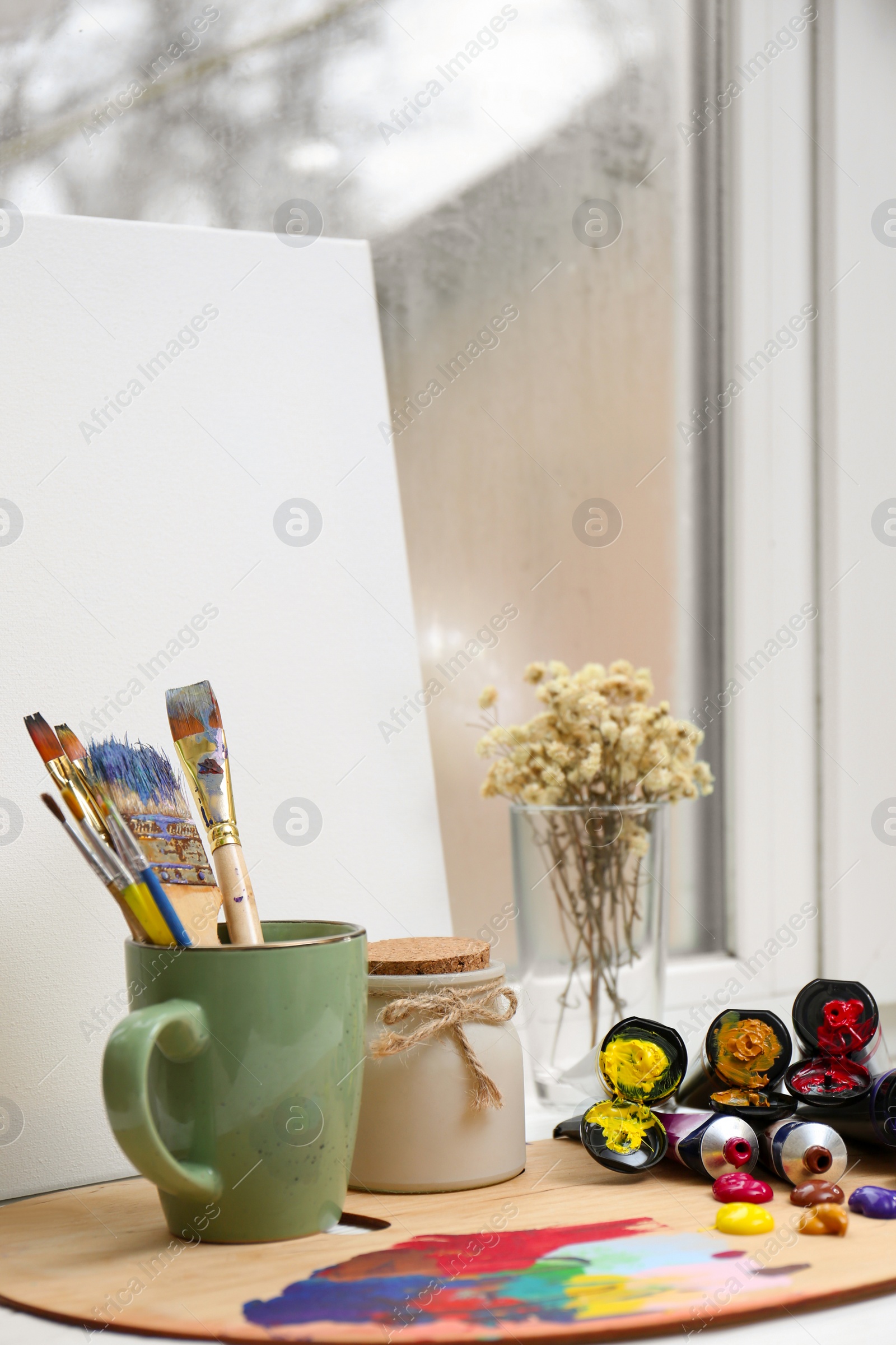 Photo of Wooden artist's palette with colorful paints, brushes and canvas on white windowsill