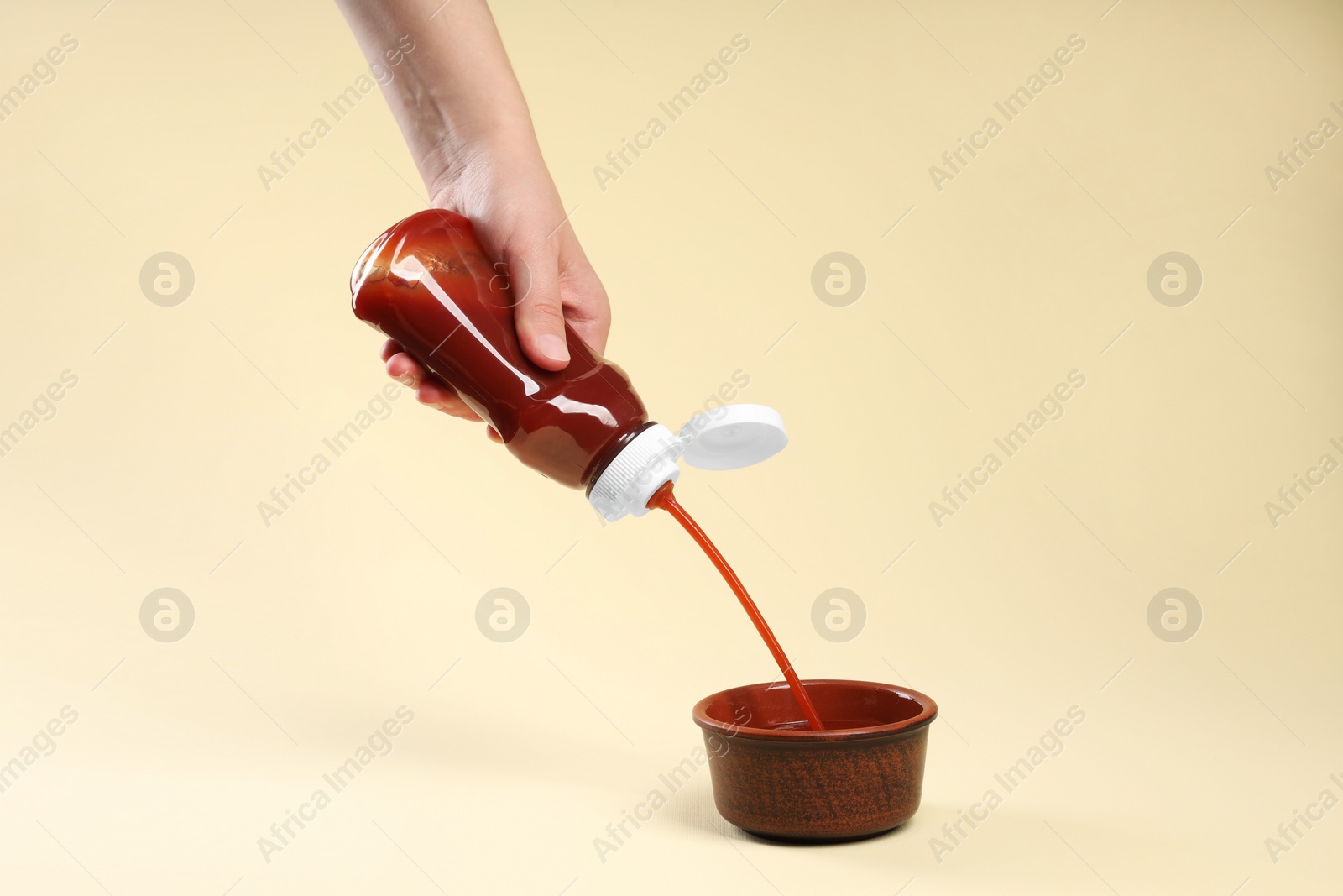 Photo of Woman pouring tasty ketchup from bottle into bowl on beige background, closeup