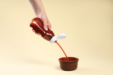 Woman pouring tasty ketchup from bottle into bowl on beige background, closeup