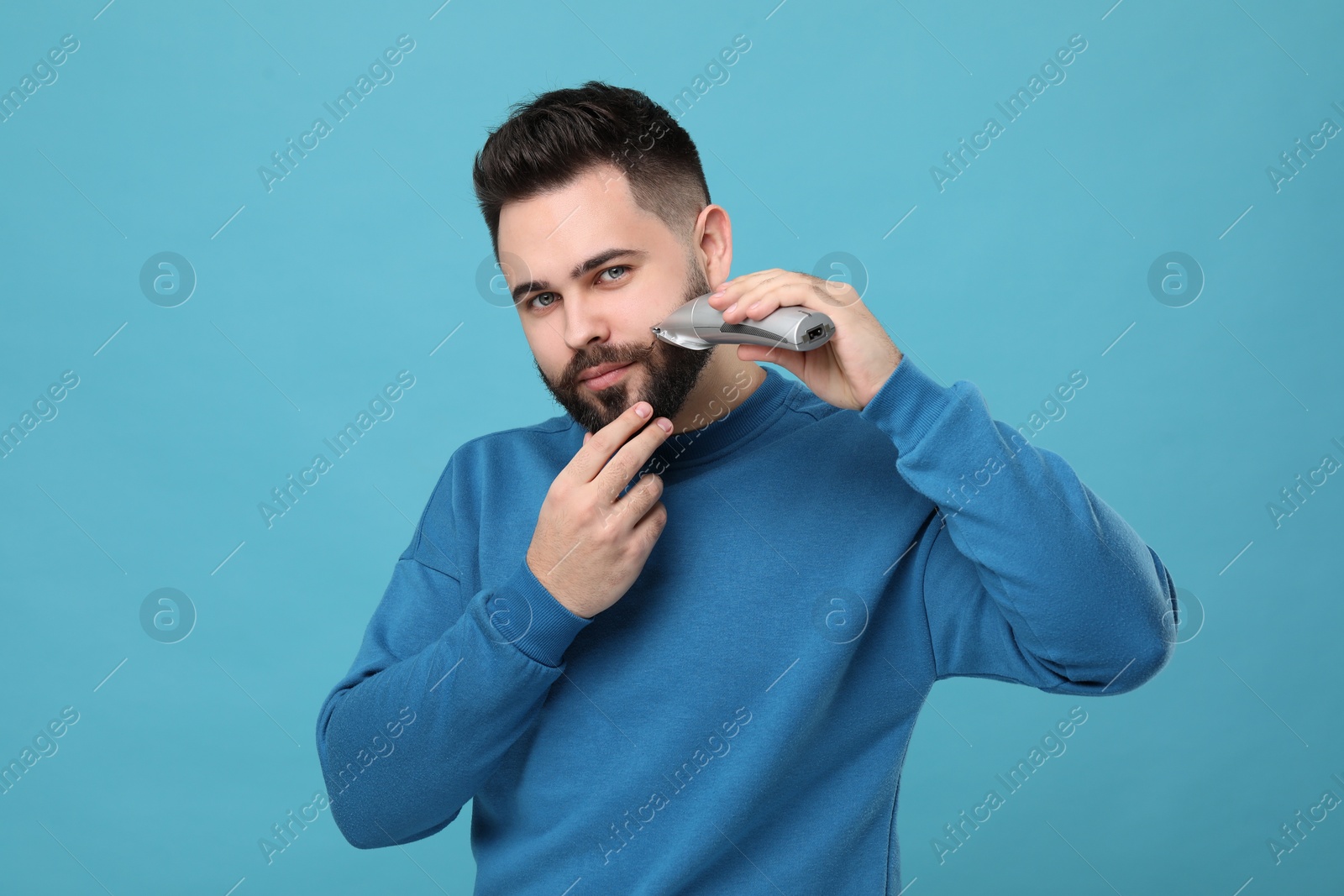 Photo of Handsome young man trimming beard on light blue background