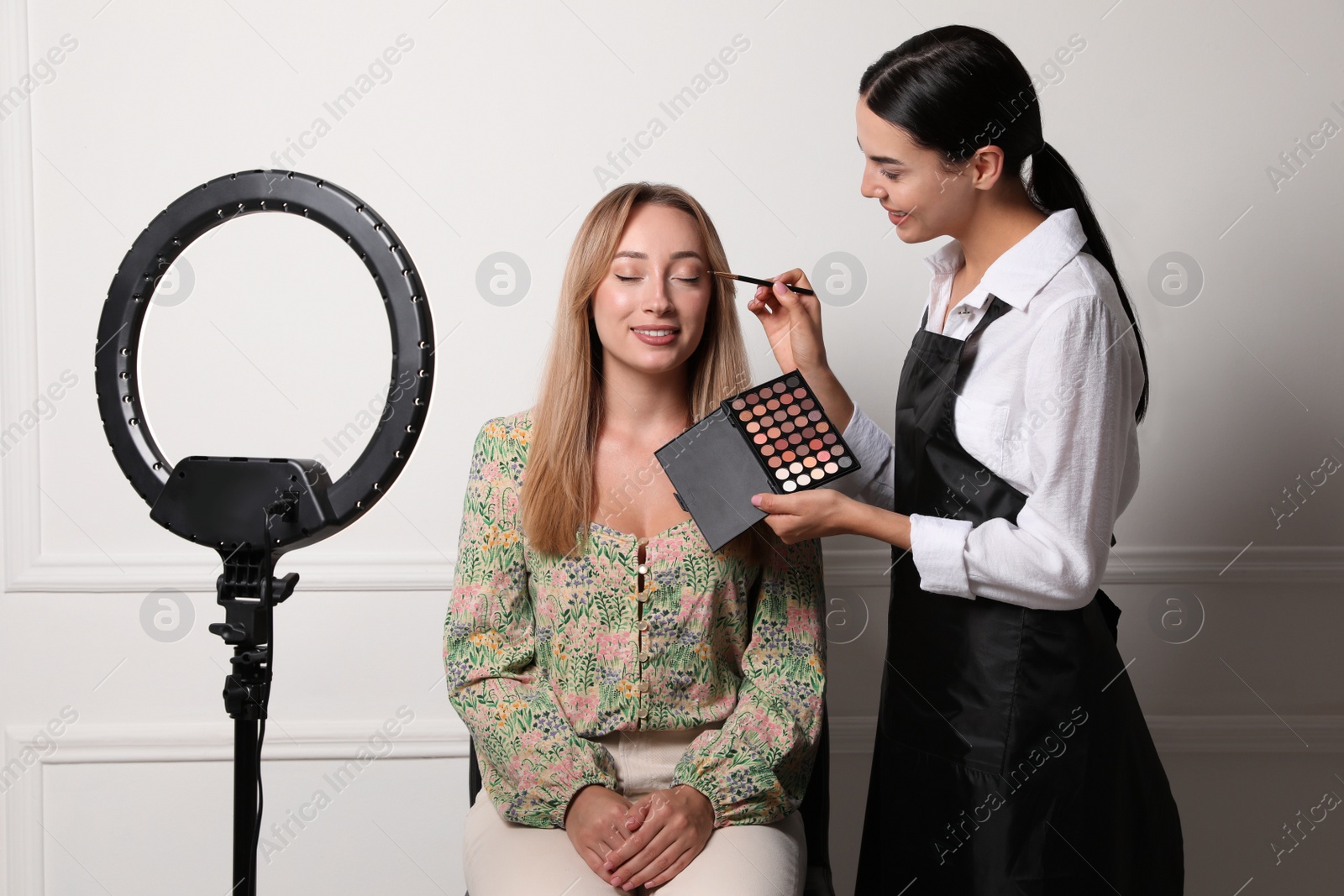 Photo of Professional makeup artist working with beautiful young woman near white wall. Using ring lamp