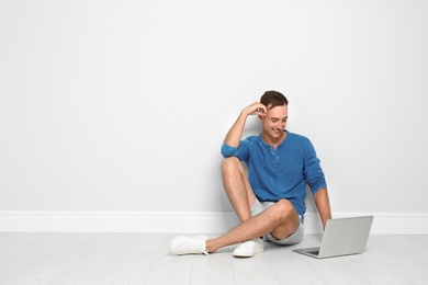 Young man with laptop sitting on floor against light wall. Space for text