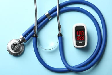Photo of Modern fingertip pulse oximeter and stethoscope on light blue background, flat lay