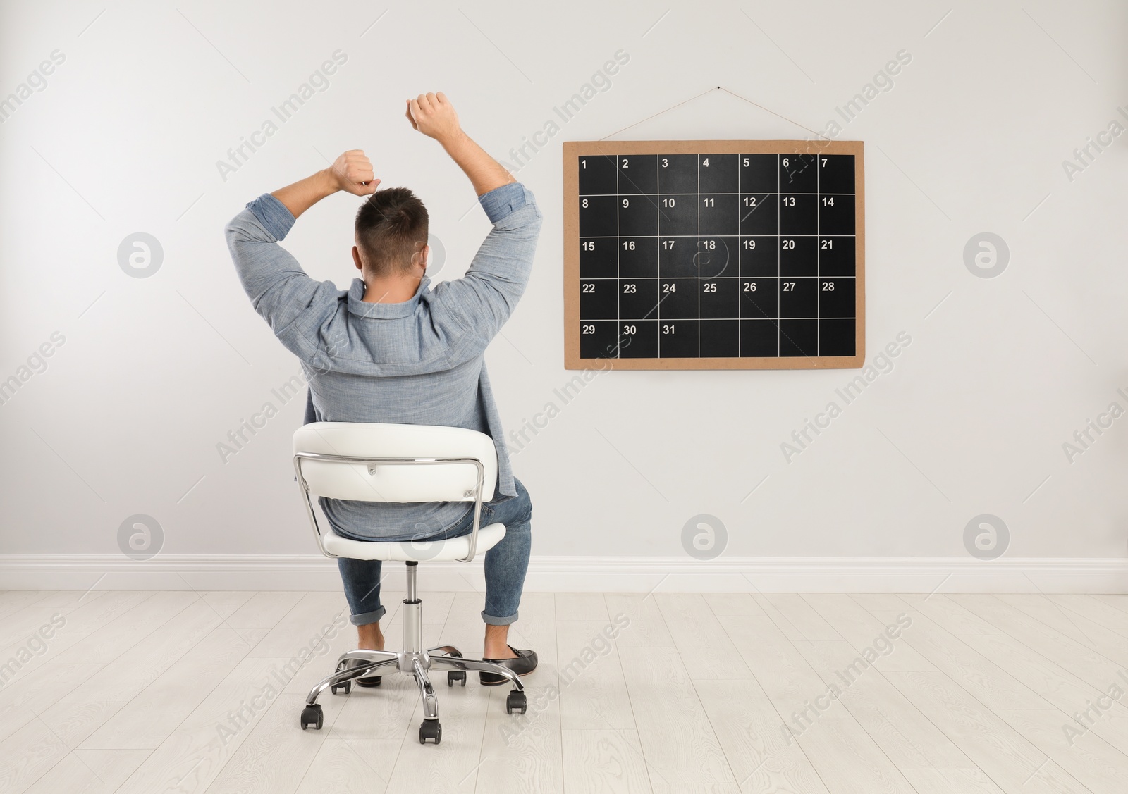 Photo of Young man sitting near board calendar indoors, back view