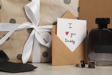 Photo of Happy Father's Day. Card with phrase I Love You, Daddy in envelope, cufflinks, perfume and gift box on light marble table, closeup
