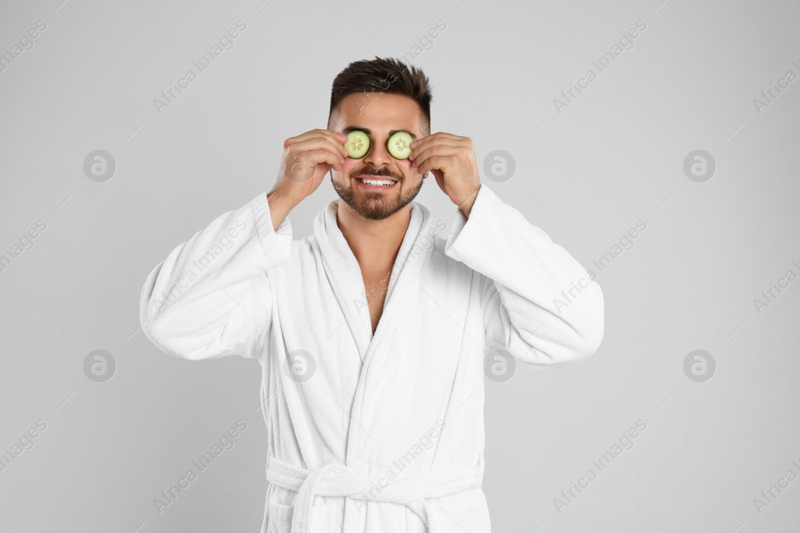 Photo of Young man in bathrobe with cucumber slices on light grey background