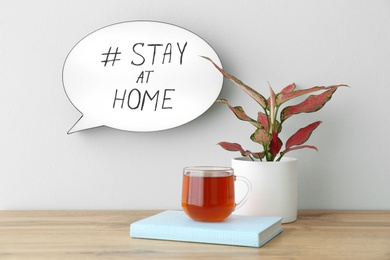 Photo of Houseplant, book, cup of tea and speech bubble with hashtag STAY AT HOME on white wall. Message to promote self-isolation during COVID‑19 pandemic