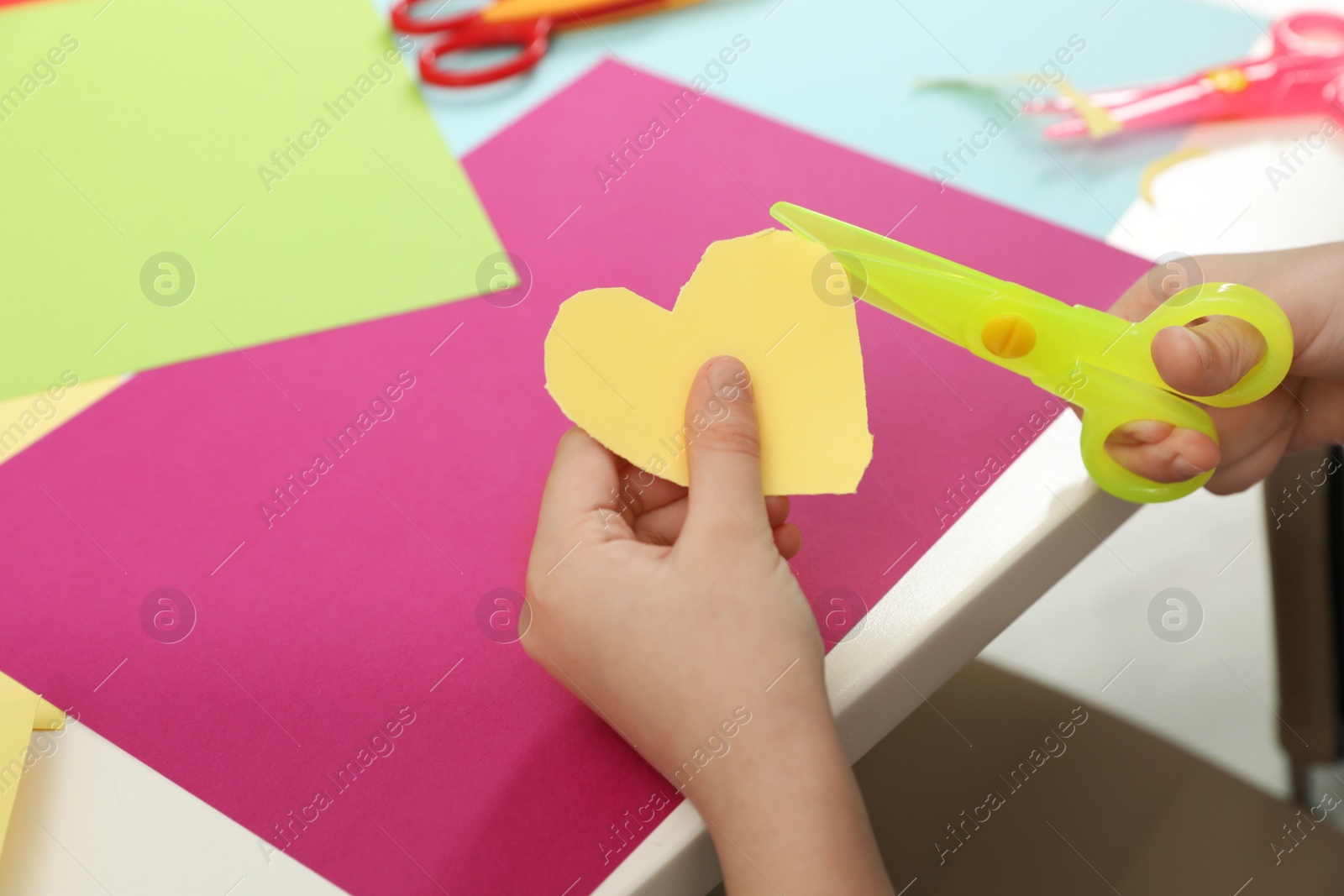 Photo of Child cutting out paper heart with plastic scissors at table, closeup. Space for text