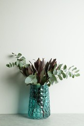 Bouquet of beautiful protea flowers and eucalyptus branches in glass vase on grey table against white background. Space for text