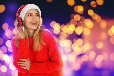 Happy woman in Santa hat listening to Christmas music with headphones on bright background, bokeh effect