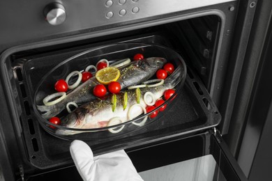 Woman putting glass baking tray with sea bass fish and ingredients into oven, closeup