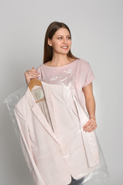 Young woman holding hanger with jacket in plastic bag on light grey background. Dry-cleaning service