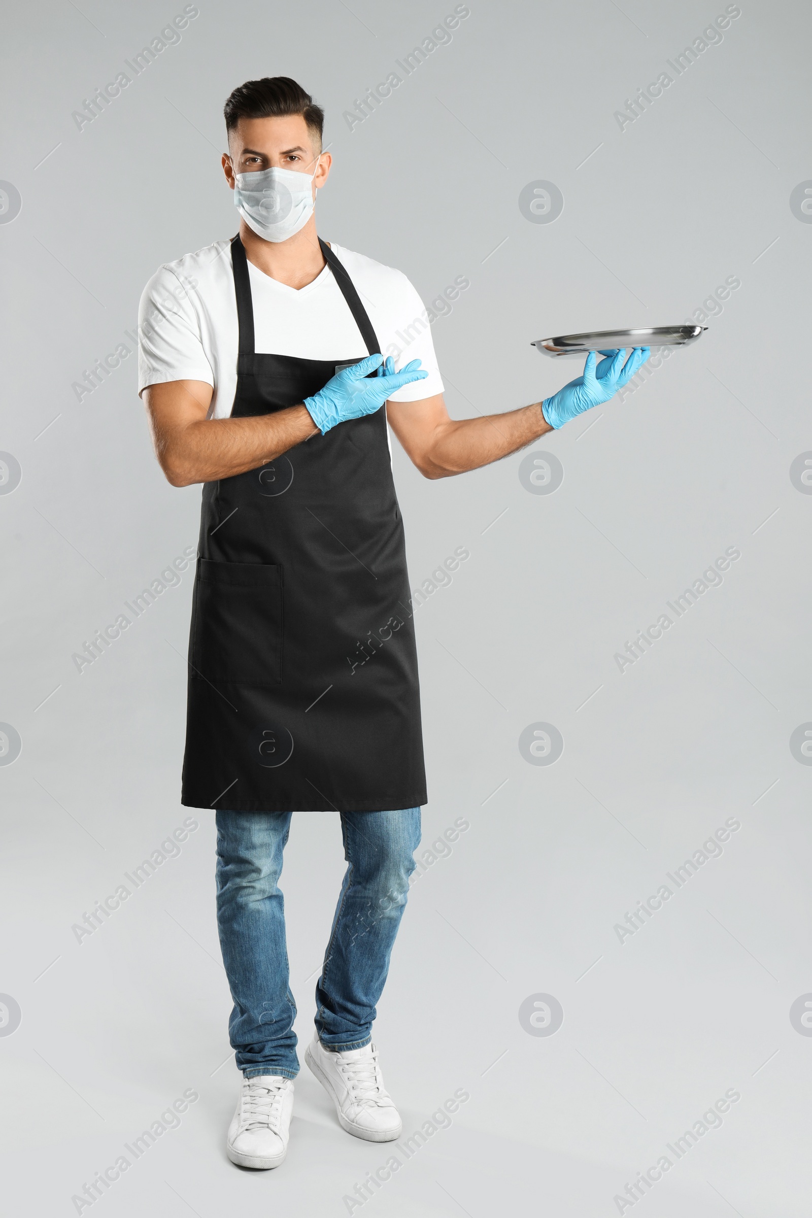 Photo of Waiter in medical face mask with empty tray on light grey background