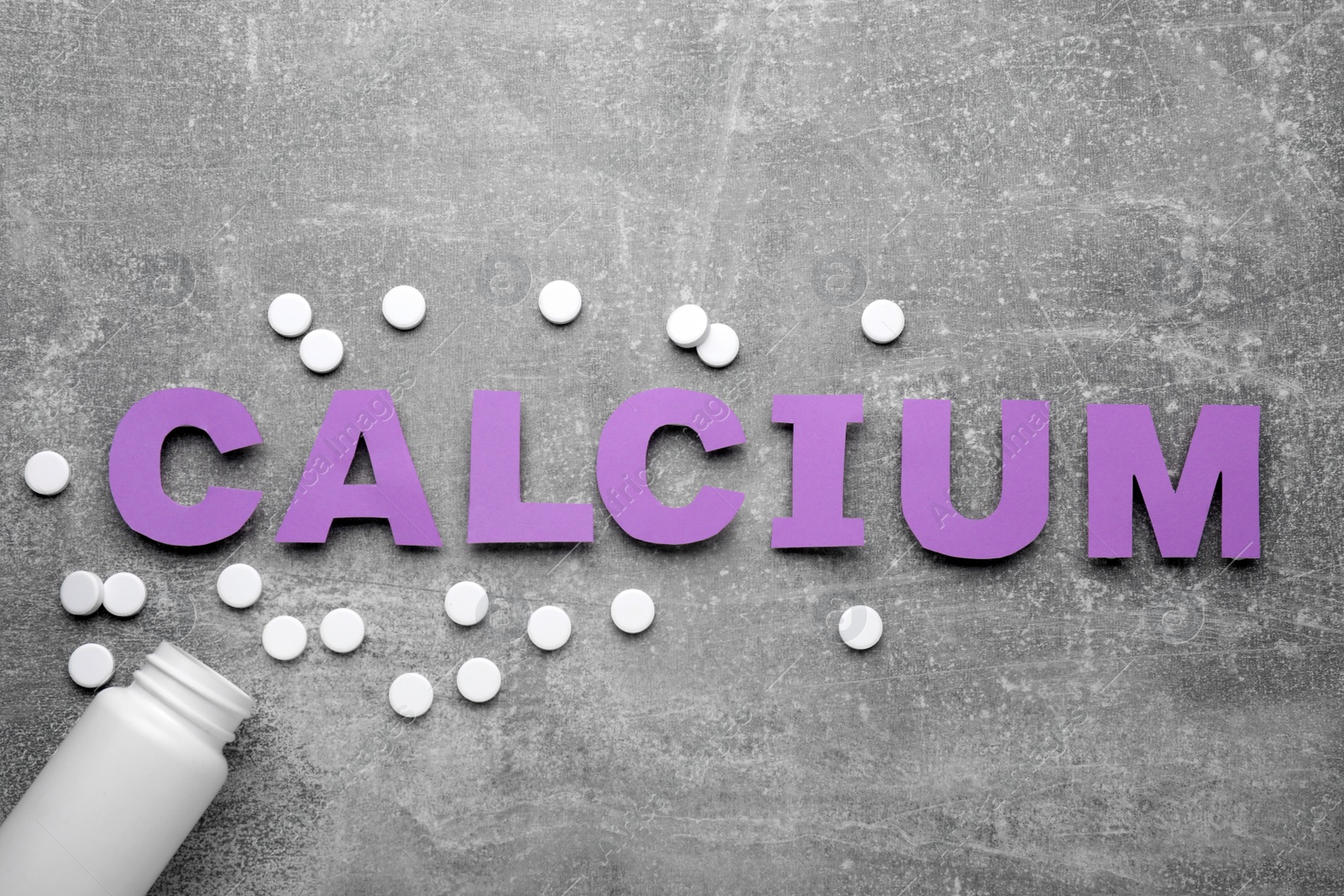 Photo of Word Calcium made of violet paper letters, medical bottle and pills on gray background, top view