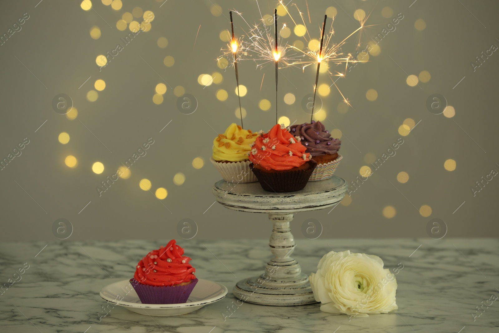 Photo of Different colorful cupcakes with sparklers on white marble table against blurred lights