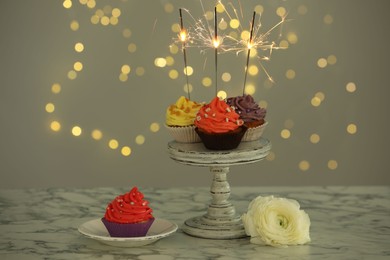 Photo of Different colorful cupcakes with sparklers on white marble table against blurred lights