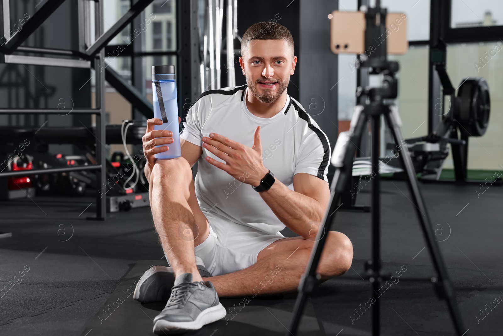 Photo of Man with bottle of water streaming online training on phone at gym. Fitness coach