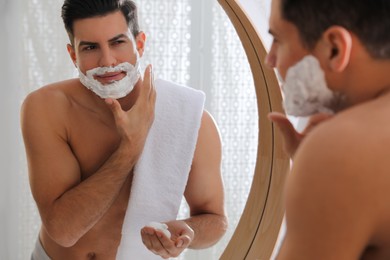 Handsome man applying shaving foam near mirror in bathroom