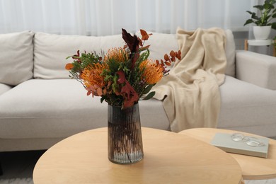 Photo of Vase with bouquet of beautiful leucospermum flowers, book and glasses on wooden nesting tables near beige sofa in room