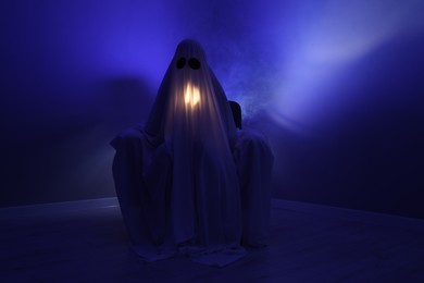 Photo of Creepy ghost. Woman covered with sheet sitting in armchair in blue light