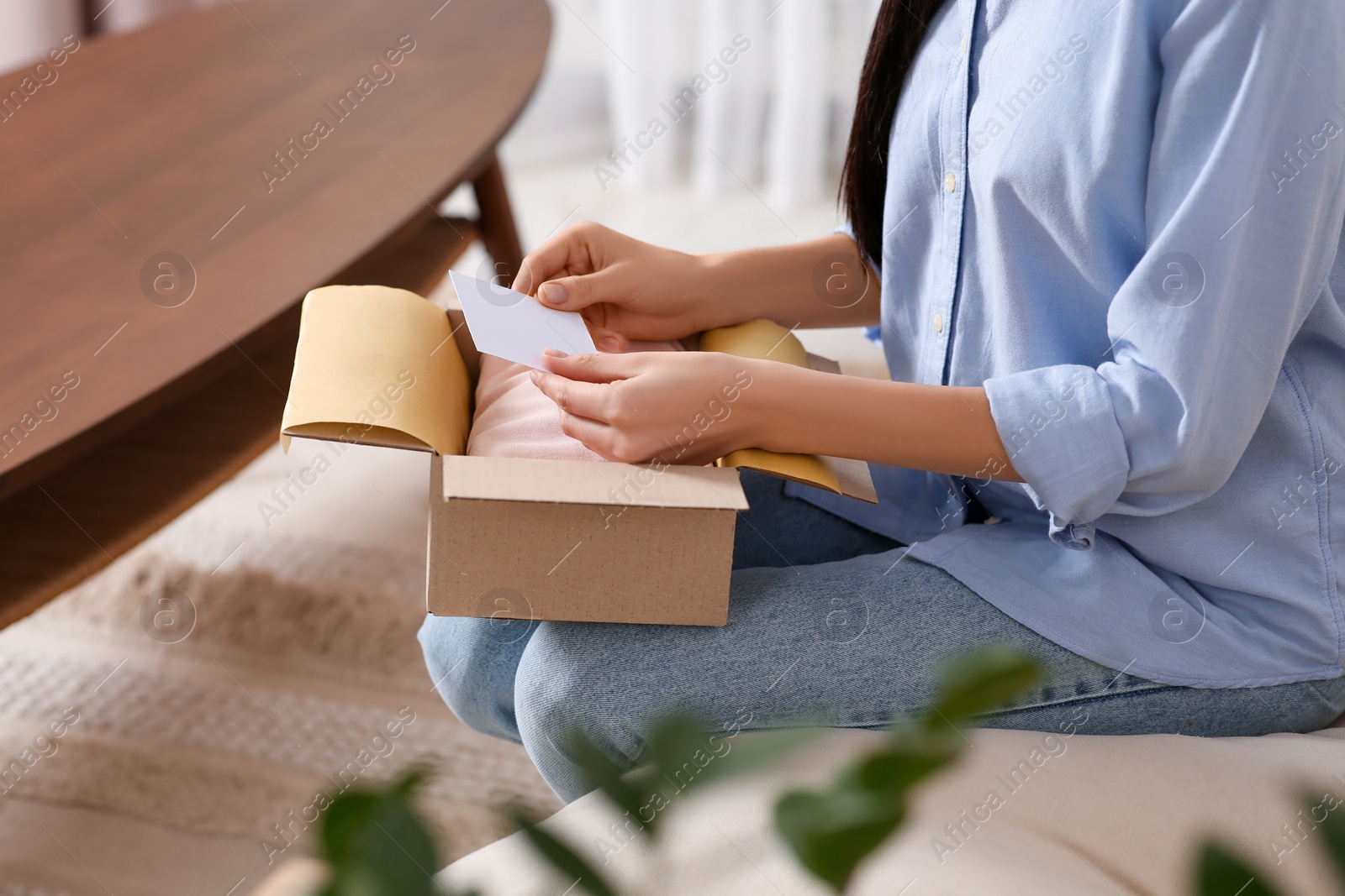 Photo of Woman holding blank greeting card near package with gift indoors, closeup