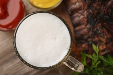 Photo of Mug with beer, delicious grilled ribs and sauces on wooden table, closeup