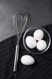 Metal whisk and raw eggs on dark grey table, flat lay