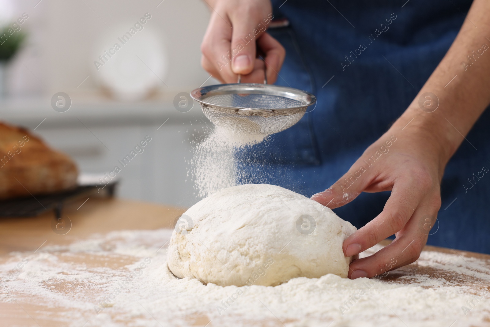 Photo of Making bread. Man sprinkling flour onto dough at wooden table in kitchen, closeup