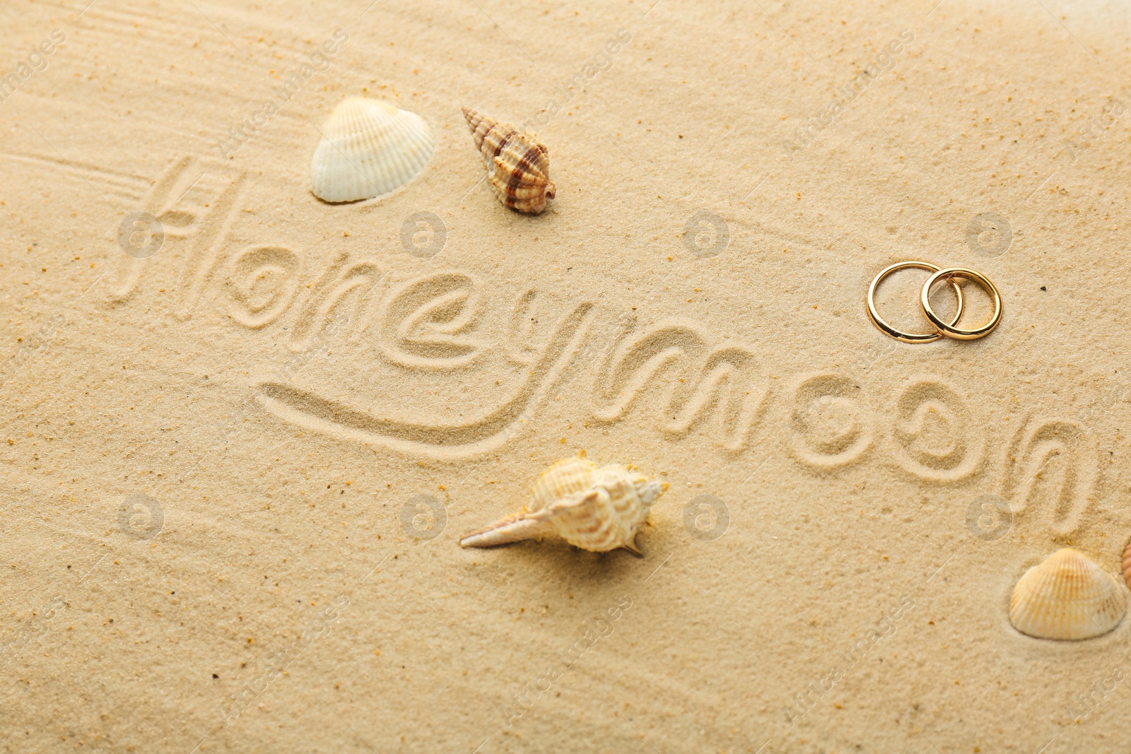 Photo of Word Honeymoon written on sand, two golden rings and seashells, closeup