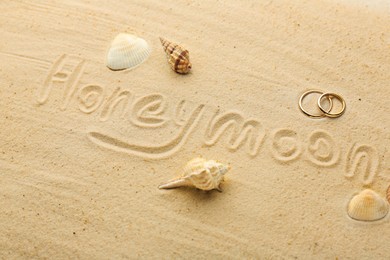 Photo of Word Honeymoon written on sand, two golden rings and seashells, closeup