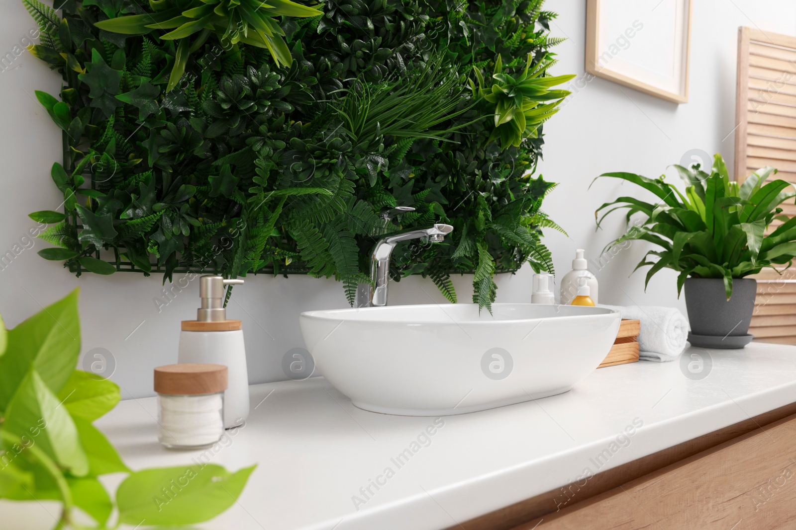 Photo of Green artificial plants, vessel sink and different personal care products in bathroom