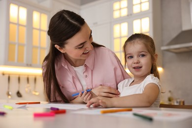 Mother and her little daughter drawing with colorful markers at table in kitchen