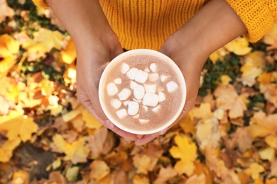 Photo of Woman holding cup of hot drink in park with fallen leaves, above view. Autumn season