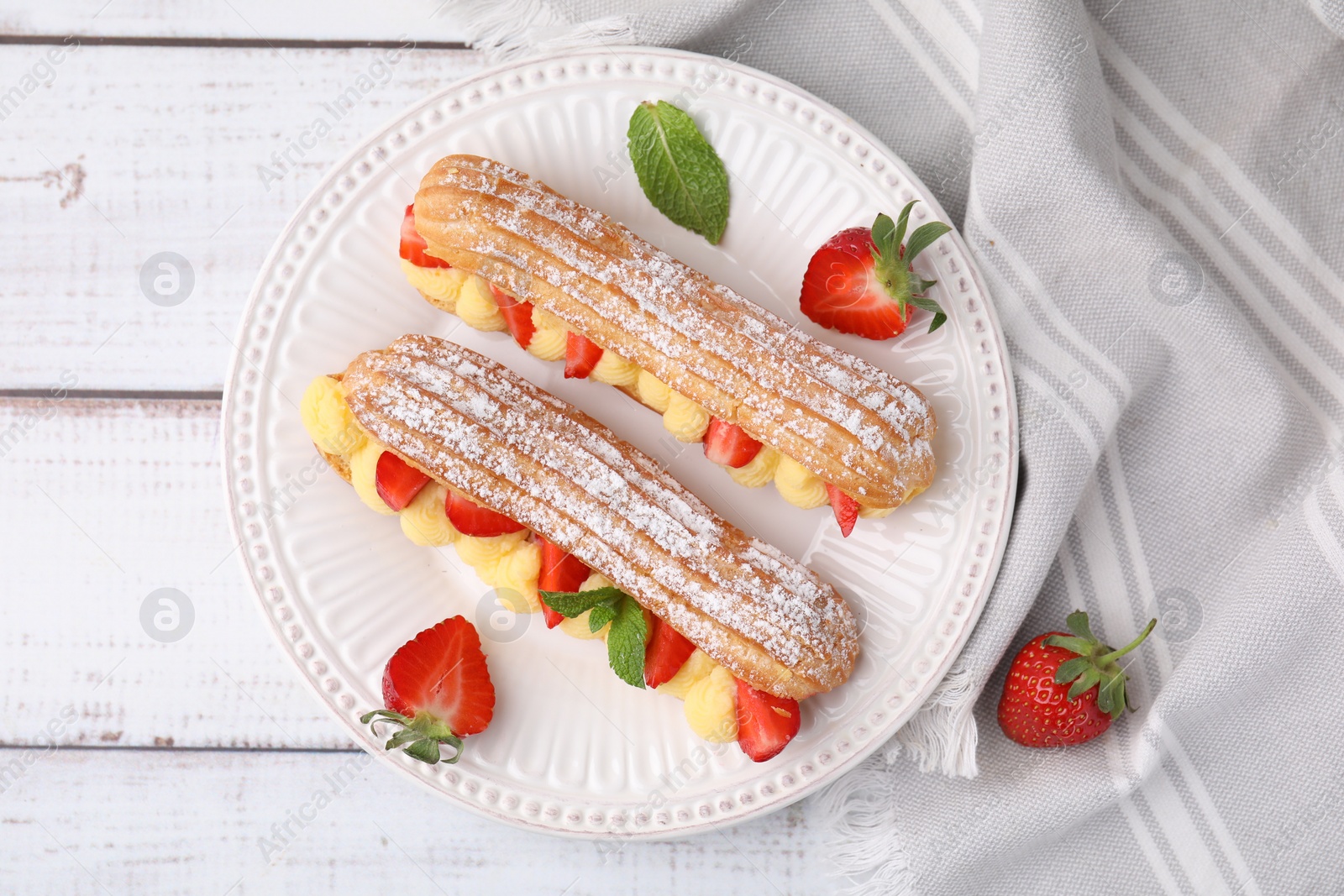 Photo of Delicious eclairs filled with cream, strawberries and mint on white wooden table, top view