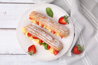 Delicious eclairs filled with cream, strawberries and mint on white wooden table, top view