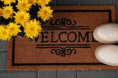 Door mat with word Welcome, stylish boots and beautiful flowers on floor, flat lay