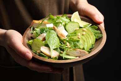 Photo of Woman holding bowl of delicious salad with chicken, arugula and avocado on dark background, closeup