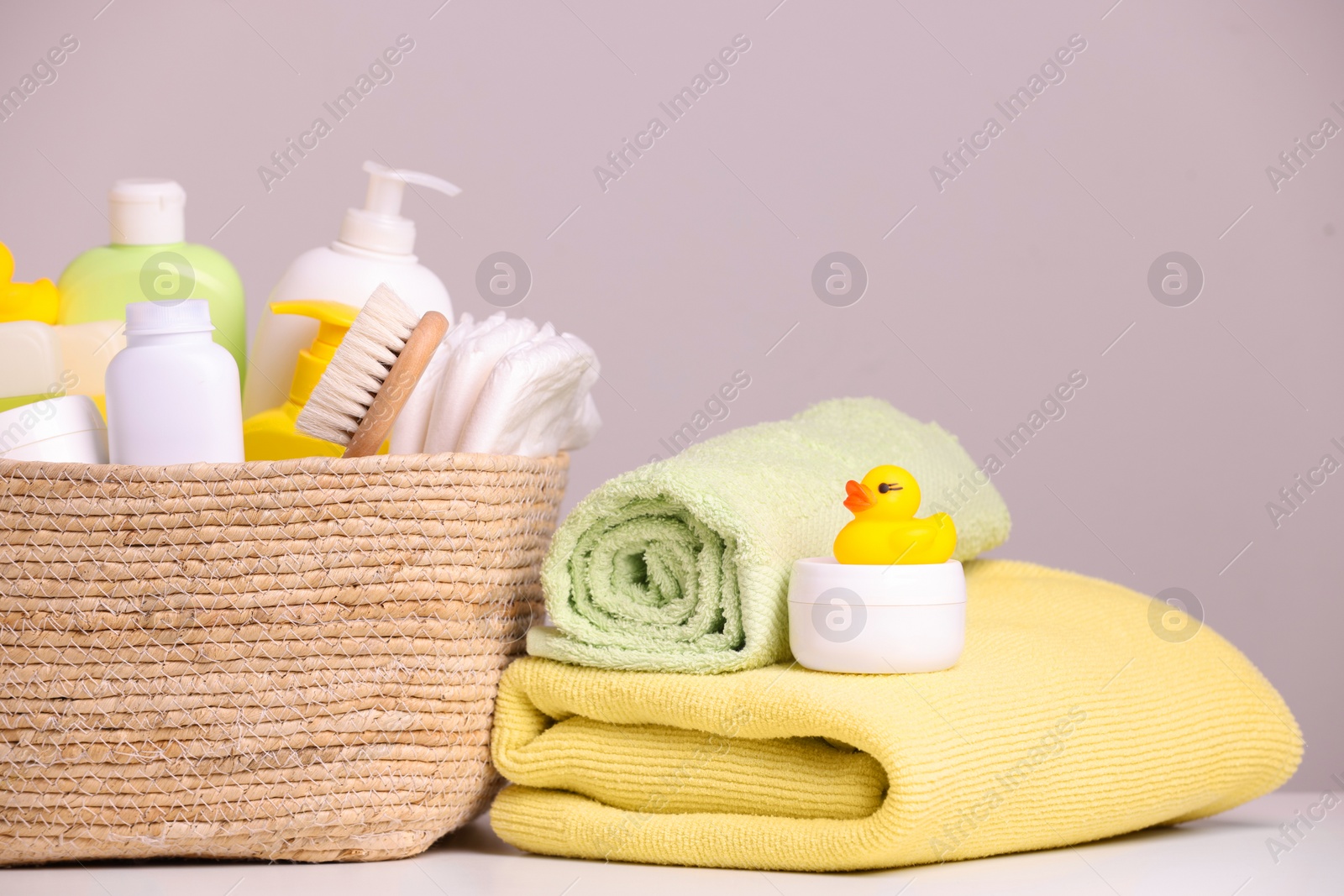 Photo of Wicker basket with baby cosmetic products, bath accessories and rubber duck on white table against grey background