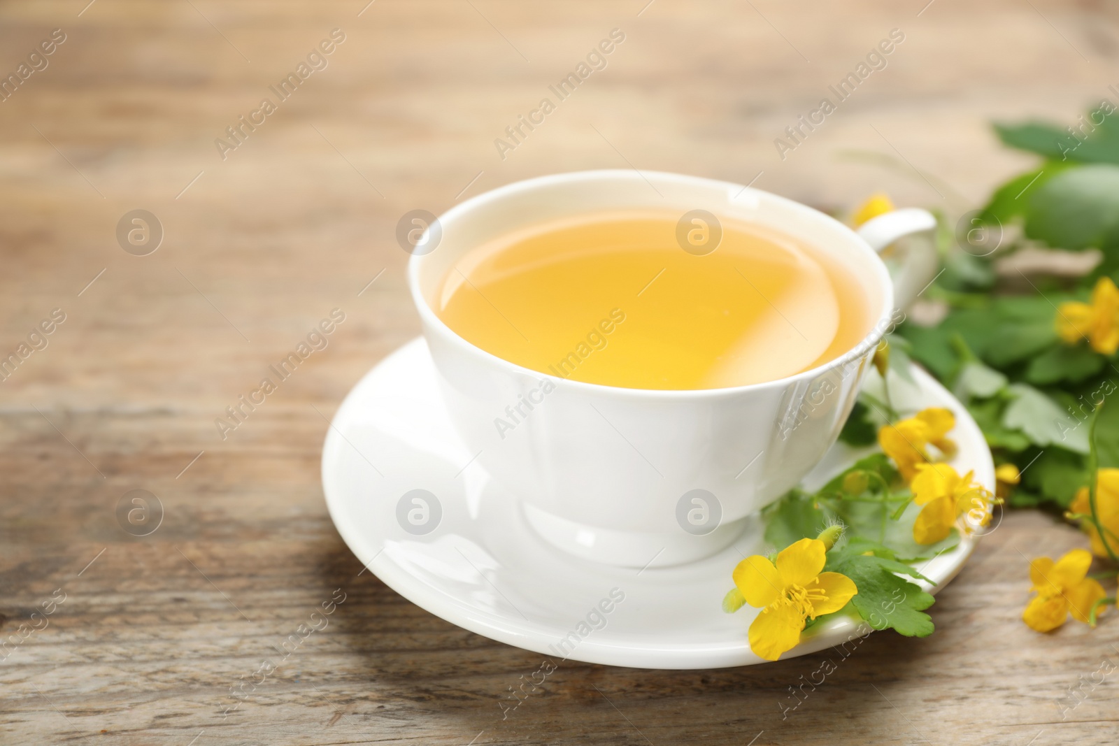 Photo of Cup of aromatic celandine tea and flowers on wooden table, closeup