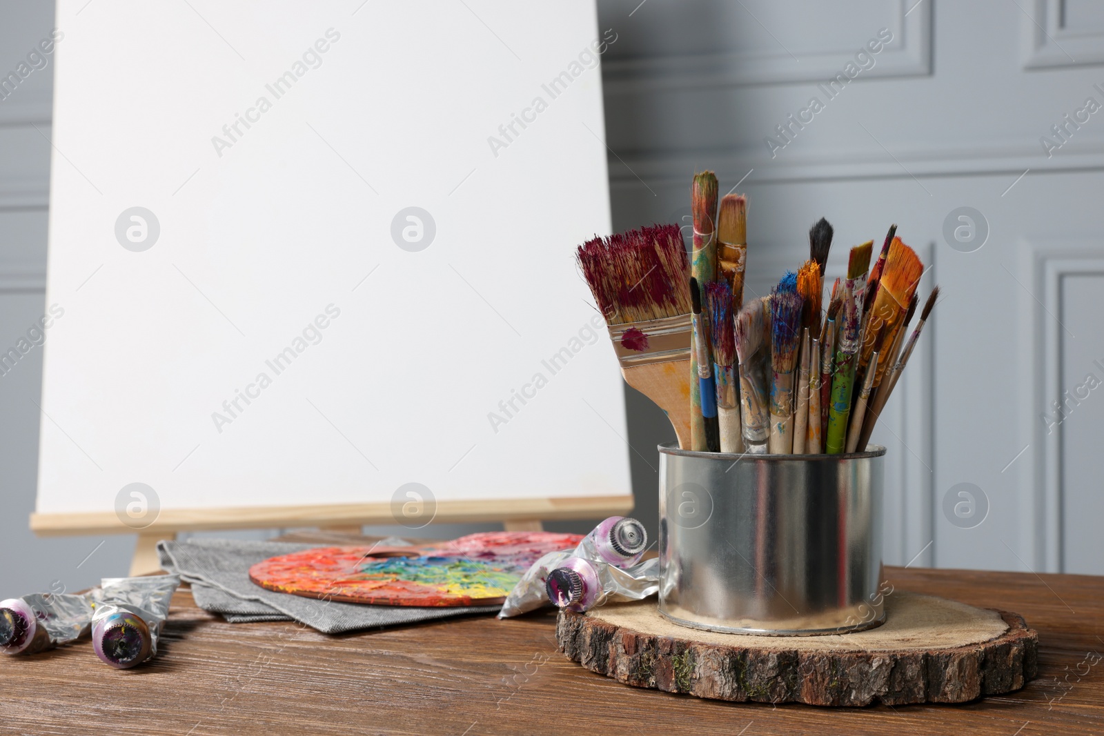Photo of Easel with blank canvas and different art supplies on wooden table near grey wall