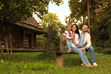 Photo of Happy cute family in embroidered Ukrainian shirts sitting on rustic wooden bench near house. Space for text