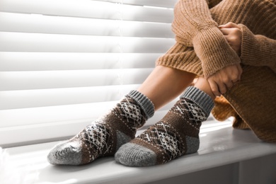 Woman wearing knitted socks on window sill indoors, closeup. Warm clothes