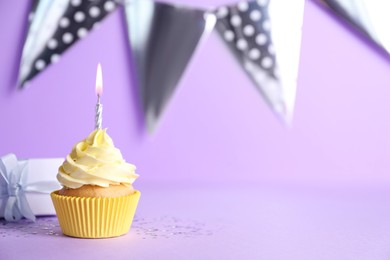 Photo of Delicious birthday cupcake with burning candle and gift box on violet background, space for text