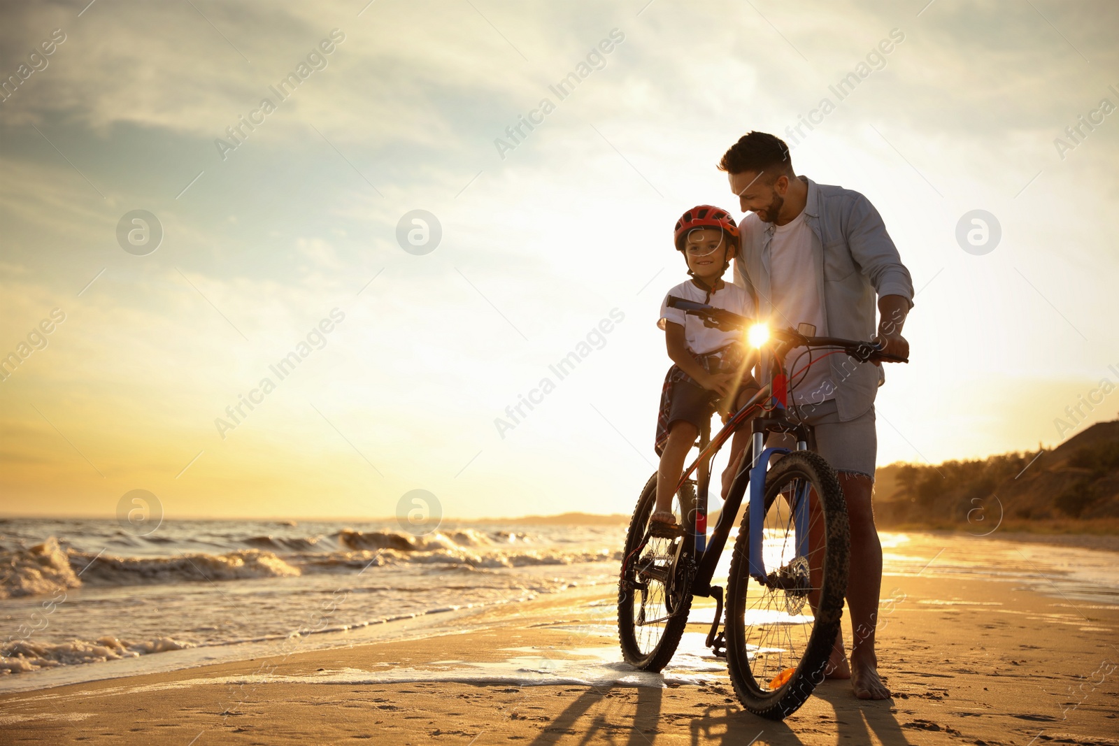 Photo of Happy father teaching son to ride bicycle on sandy beach near sea at sunset