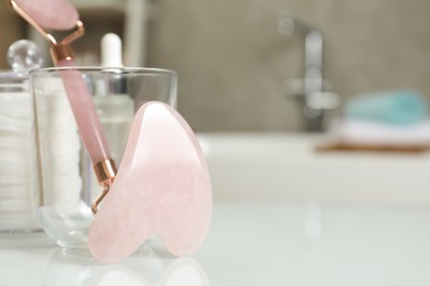 Photo of Rose quartz gua sha tool, natural face roller and toiletries on white table in bathroom, closeup. Space for text